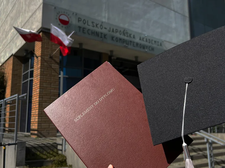 The diploma supplement held up in front of the PJAIT building in Warsaw, a graduation cap beside it